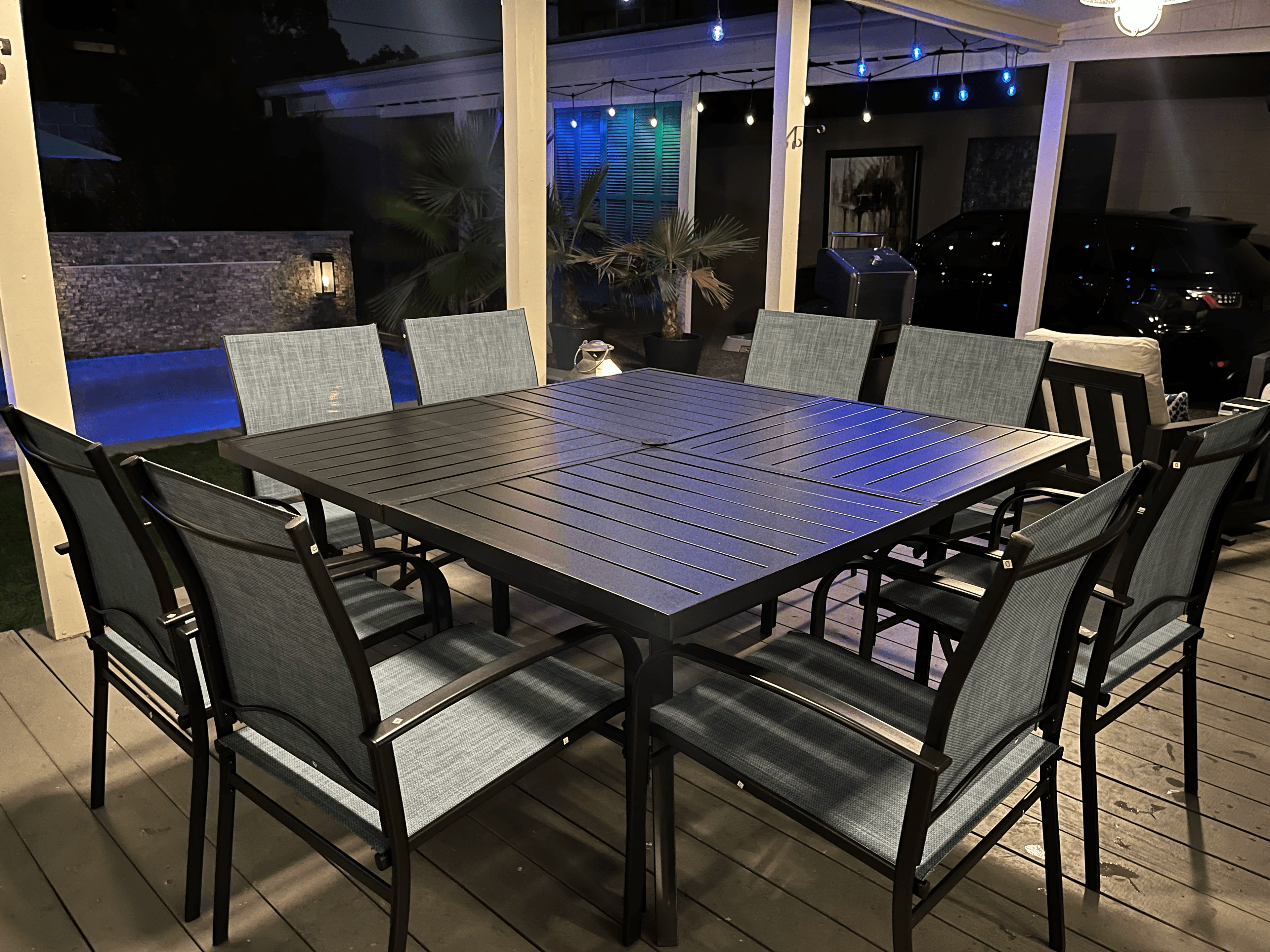 Square dining table and eight chairs on a deck at night near a lit pool.