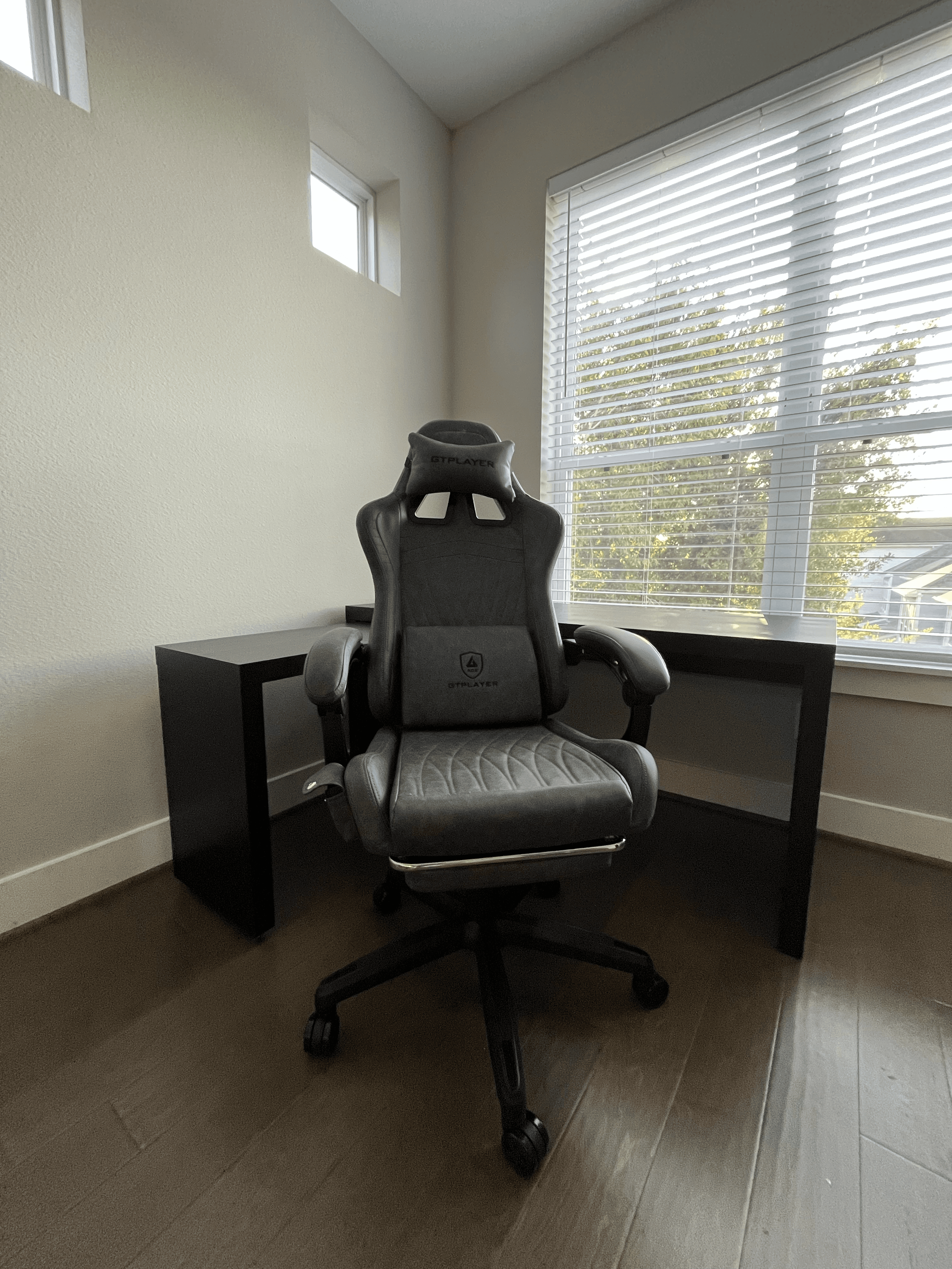 Grey GTPlayer gaming chair in front of a black desk near a window with blinds.